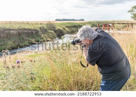 Photographer with gray hair taking pictures with camera of thistles in the field near the river.