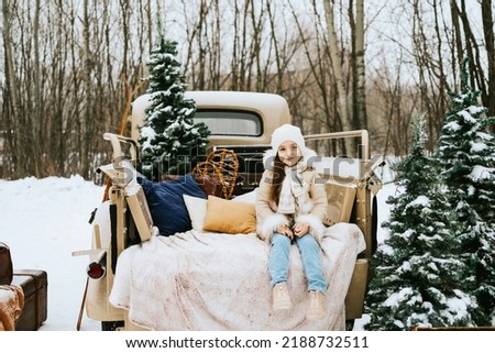 cute beautiful little Armenian girl in winter coat and knitted hat sit near beige retro pickup truck decorated Christmas and New Year vintage interior items, suitcases, skis, sledge, ornaments, travel
