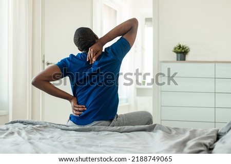 Rear View Of African American Man Touching And Massaging Aching Neck And Spine Suffering From Pain After Sleeping On Uncomfortable Bed Sitting In Modern Bedroom At Home. Arthritis Symptom Royalty-Free Stock Photo #2188749065