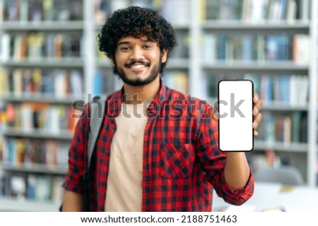 Gadgets, mock-up concept. Defocused positive attractive indian or arabian guy, freelancer or student, showing his smartphone with blank white mock-up screen, looks at camera, smiles happily