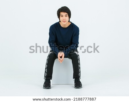 Studio full body shot of Asian young handsome confident slim healthy athletic teenager fashion male model in long sleeve shirt fabric pants and sneaker sitting on box posing on white background.