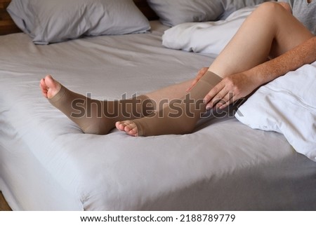 Woman sitting on bed putting on compression socks on her legs Royalty-Free Stock Photo #2188789779