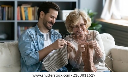 Happy mature mother and son knitting together, enjoying leisure time at home, smiling middle aged woman wearing glasses and young man sitting on cozy couch in living room, holding needles Royalty-Free Stock Photo #2188811629