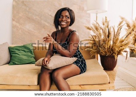 Happy stylish beautiful female student with hair looks at camera stands in cozy home interior. Smiling attractive young woman wears black dress headshot portrait.