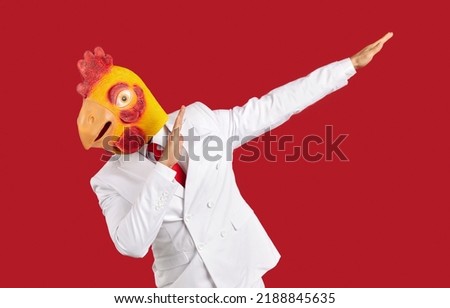 Weirdo man in chicken mask makes movement of famous internet meme about victory on red background. Young man in white suit and with rubber head mask makes dabbing movement during costume party