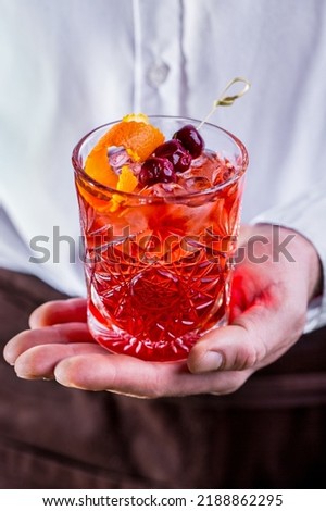 A cocktail with ice, cherries on a skewer and an orange peel in a glass held by a man against the background of a black and white wall