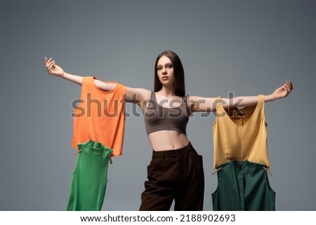 pretty model in crop top and trousers standing with outstretched hands and holding clothing on grey