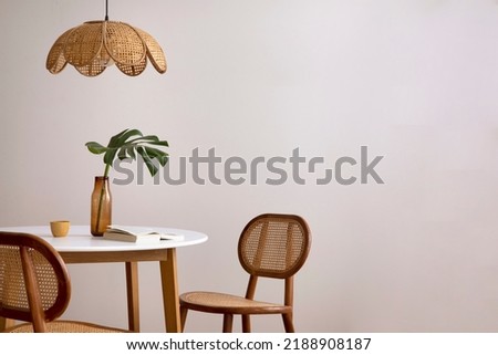 Minimalist composition of elegant kitchen space with round table, rattan chair, vase with leaf, pedant lamp and personal accessories. Minimalist home decor. Beige wall. Template.  Royalty-Free Stock Photo #2188908187