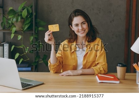Young smiling happy successful employee business woman 20s wear casual yellow shirt hold in hand mock up of credit bank card sit work at wooden office desk with pc laptop. Achievement career concept Royalty-Free Stock Photo #2188915377