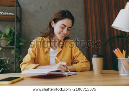 Young smiling happy cheerful fun european successful employee business woman 20s she wearing casual yellow shirt writing in notebook diary sit work at wooden office desk. Achievement career concept Royalty-Free Stock Photo #2188915395