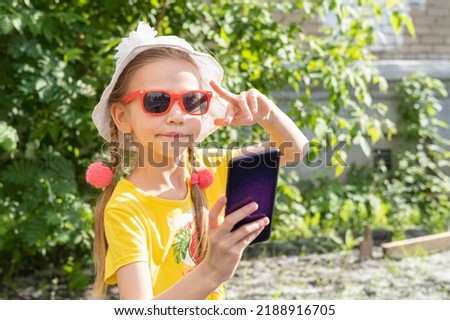 Portrait of little girl child taking a selfie by smartphone in summer park. A cheerful little girl in a yellow dress, white hat and sunglasses takes a photo on her phone and shows a V gesture.
