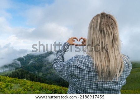 Young blonde woman in a plaid shirt is in the mountains at the level of clouds and makes a love heart shape with her hands, photo from the back