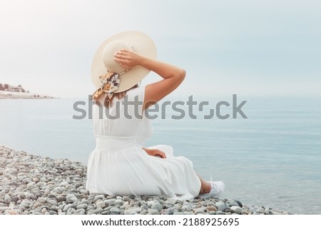 A young girl in a white dress is sitting on the seashore in the rays of the sun, looking into the distance. She holds the straw hat with her hand.