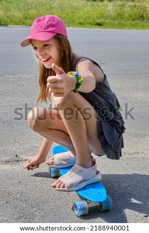 Portrait of a Caucasian smiling girl with long hair in a pink baseball cap, sitting on a skateboard on an asphalt road in sunny weather. A child rides a skateboard