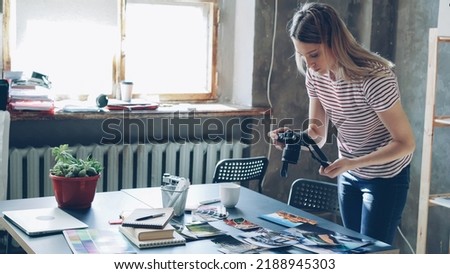 Young attractive female blogger using digital camera to shoot colourful pictures from travel displayed on table. She is moving photos and making different collages. Loft style office in background.
