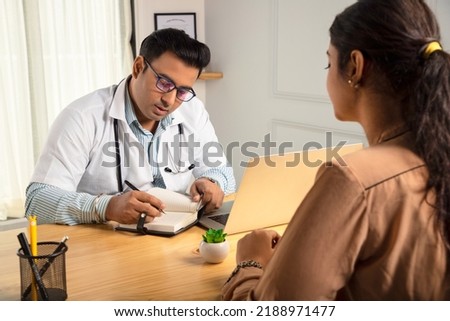 An Indian man or male physician or doctor sitting in a modern clinic wearing a stethoscope and apron writing a drug prescription to a female patient customer. Medical, medicine and healthcare concept Royalty-Free Stock Photo #2188971477