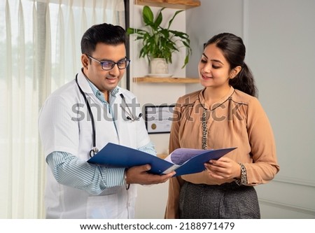 Asian Indian man or male physician or doctor wearing stethoscope and apron holding report file in hand and giving consultation to a smiling female or happy Woman patient. Medical, medicine, healthcare Royalty-Free Stock Photo #2188971479