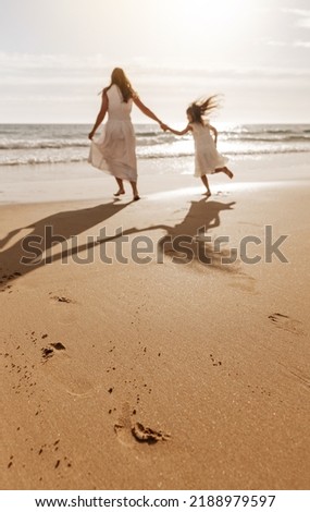 Back view of unrecognizable young woman with long dark hair in maxi white dress, holding hand of daughter while running on sandy beach towards wavy ocean at sunset