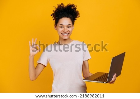 Happy pretty, curly haired african american young woman, in casual clothes, holding an open laptop in her hand, looks at camera, showing OKAY gesture, agree sign, stands on isolated orange