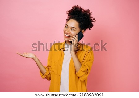 Cell phone conversation concept. Happy excited curly haired african american young woman, talking on smartphone, standing over isolated pink background, looking away, smile friendly, gesturing hand