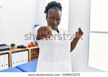 Young african woman holding covid record card at clinic pointing with finger to the camera and to you, confident gesture looking serious 