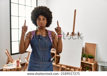 Young african american woman with afro hair at art studio pointing up looking sad and upset, indicating direction with fingers, unhappy and depressed. 