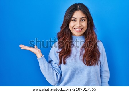 Hispanic young woman standing over blue background smiling cheerful presenting and pointing with palm of hand looking at the camera. 