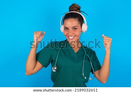 Emotional beautiful doctor woman wearing medical uniform over blue background exclaims loudly feels like winner raises clenched fists keeps mouth opened wears stereo headphones on ears makes yes gesture