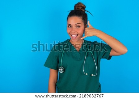 beautiful doctor woman wearing medical uniform over blue background makes phone gesture, says call me back again, has glad expression.