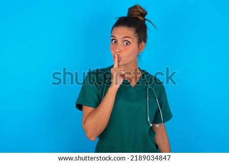 Surprised beautiful doctor woman wearing medical uniform over blue background makes silence gesture, keeps finger over lips and looks mysterious at camera