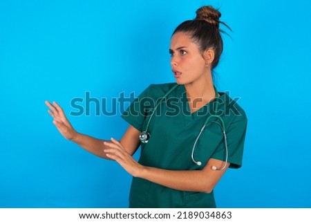 Displeased beautiful doctor woman wearing medical uniform over blue background keeps hands towards empty space and asks not come closer sees something unpleasant