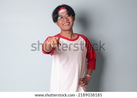 Portrait of attractive Asian man in t-shirt with red and white ribbon on head, pointing forward at the camera. Isolated image on gray background