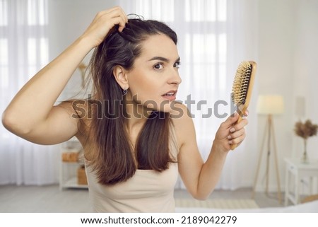 Portrait of woman in her 20s or 30s looking at her reflection with scared nervous expression as she notices bad signs like scalp dandruff, hair thinning, or hair falling out. Hair loss problem Royalty-Free Stock Photo #2189042279