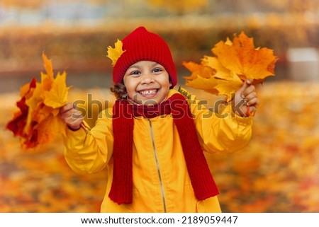 A girl in a yellow raincoat jacket, a red hat and a scarf in an autumn park holds red and yellow leaves in her hands.