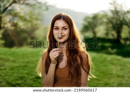 Portrait of a young woman with a dandelion flower in her hand blowing on it and smiling against the green summer grass in the rays of the setting sun in nature Royalty-Free Stock Photo #2189060617
