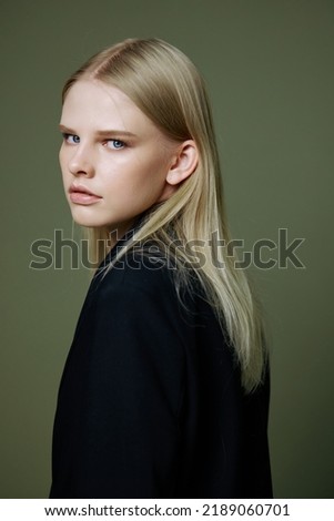 A close portrait of a blonde looking over her shoulder in a black jacket posing in a studio on a green background. The concept of stylish fashion photography Royalty-Free Stock Photo #2189060701