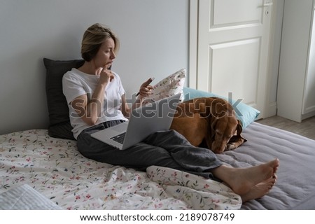 Middle-aged decisive woman calls potential employer on phone sitting on cozy bed with laptop near dog. Unemployed mature blonde woman waits for important call back looking at smartphone screen