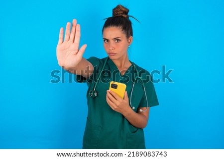 beautiful doctor woman wearing medical uniform over blue background using and texting with smartphone with open hand doing stop sign with serious and confident expression, defense gesture