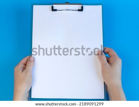 Clipboard with blank white sheet on blue background. View from above. Female hands holding clipboard, place for text.