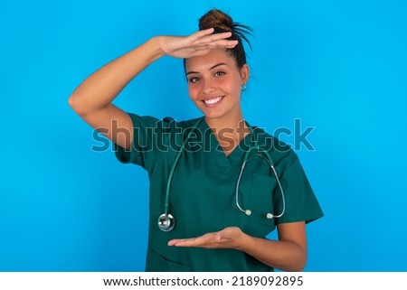beautiful doctor woman wearing medical uniform over blue background gesturing with hands showing big and large size sign, measure symbol. Smiling looking at the camera.