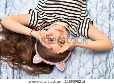 Happy relaxed little girl listening music through headphone while lying on the bed at home