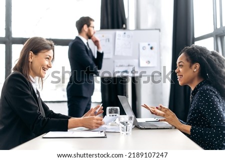 Two successful confident women are negotiating, discussing business project, planning financial strategy, smile. Elegant man stands on the background near white board, thinking business plan