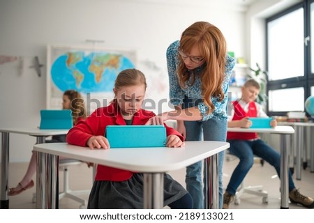 Down syndrome schoolgirl using tablet with help of teacher during class at school, integration concept. Royalty-Free Stock Photo #2189133021