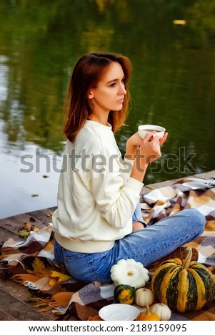A young woman drinks tea from a white mug on a wooden bridge on the lake. Rest in the warm autumn in nature.