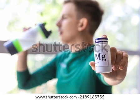 Young man with bottle of magnesium pills drinking water near window, closeup Royalty-Free Stock Photo #2189165755