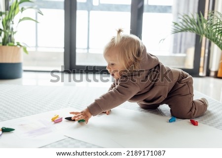 Cute caucasian baby drawing on floor in living room with potted green plant at home. Happy girl painting with crayons. 