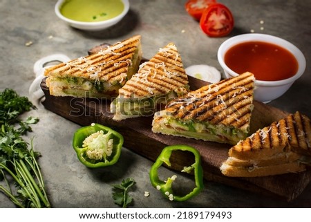 Vegetable Toste sandwich is a type of veg sandwich consisting of a vegetable filling between bread. photo is composited on grey background and wood platter with onion, capsicums  tomato, green sauces. Royalty-Free Stock Photo #2189179493