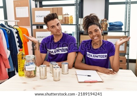 Young interracial people wearing volunteer t shirt at donations stand smiling cheerful presenting and pointing with palm of hand looking at the camera. 