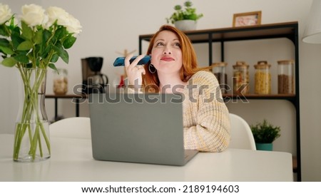 Young redhead woman using laptop listening audio message by smartphone at home