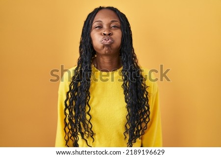 African woman standing over yellow background puffing cheeks with funny face. mouth inflated with air, crazy expression. 